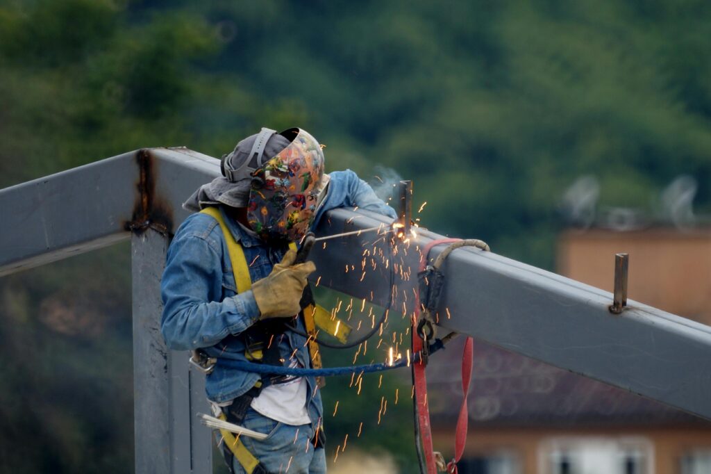 A construction worker welding a steel beam outdoors, emitting sparks, wearing safety gear.